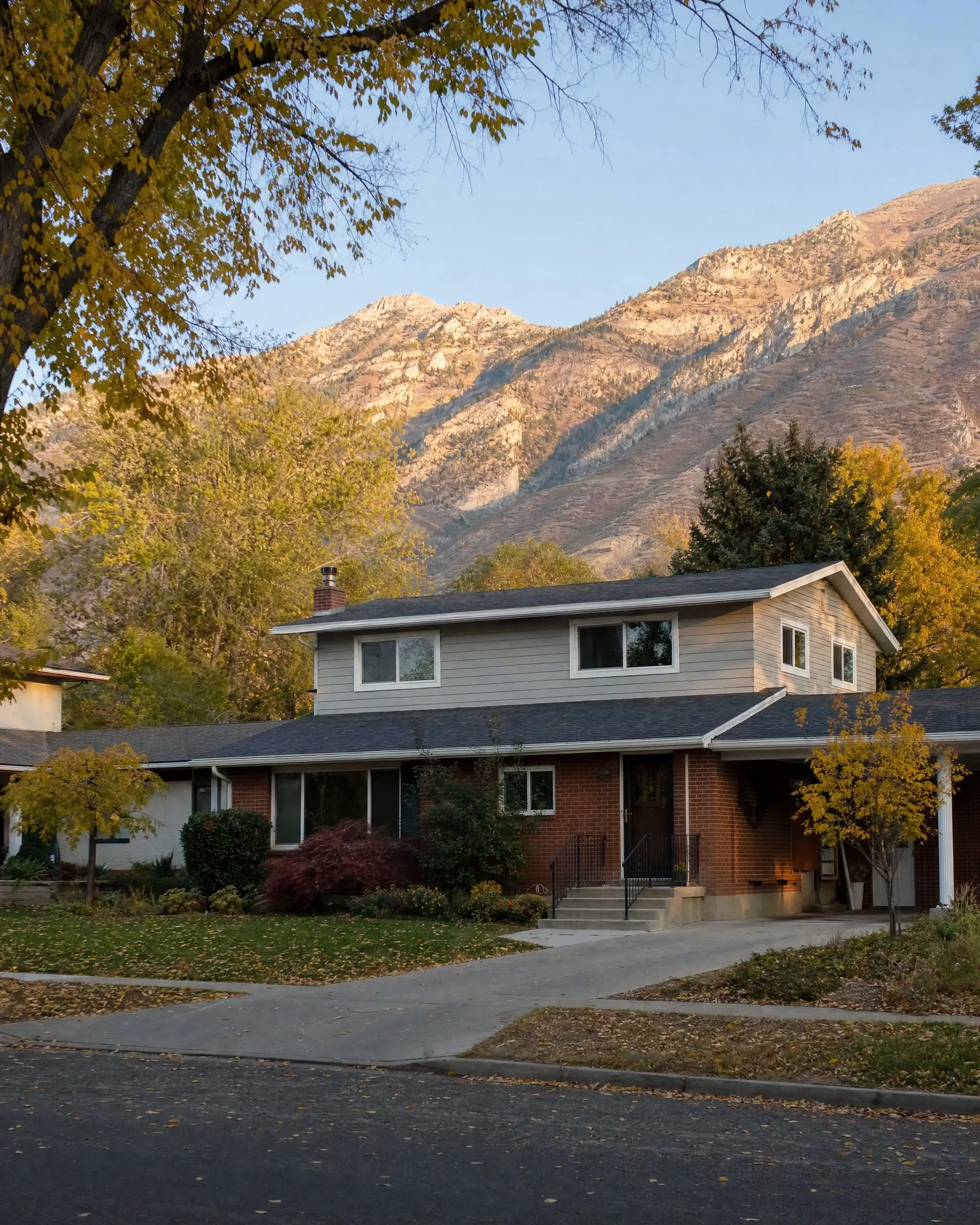 Tree-lined Bountiful street with a brick rambler that has a matching second-story addition, Wasatch mountains behind
