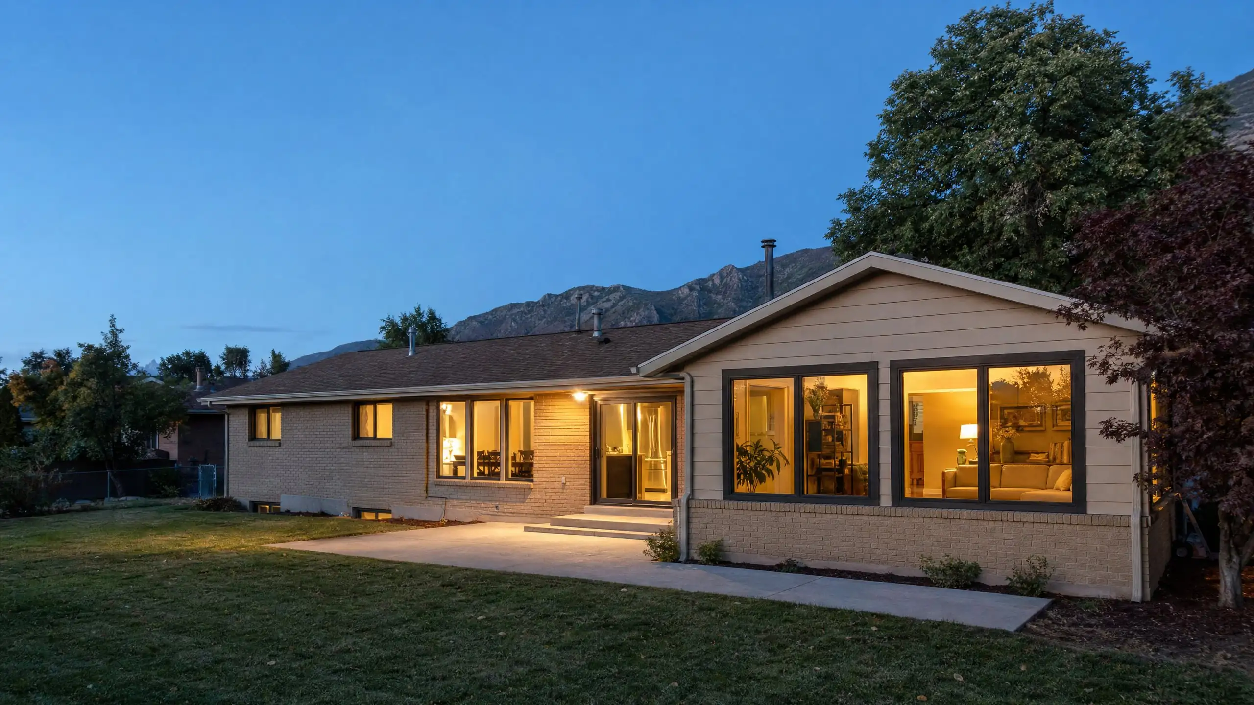 Brick rambler at dusk with a family room addition that continues the original roofline, brick and siding, its windows lit from inside, Wasatch foothills behind